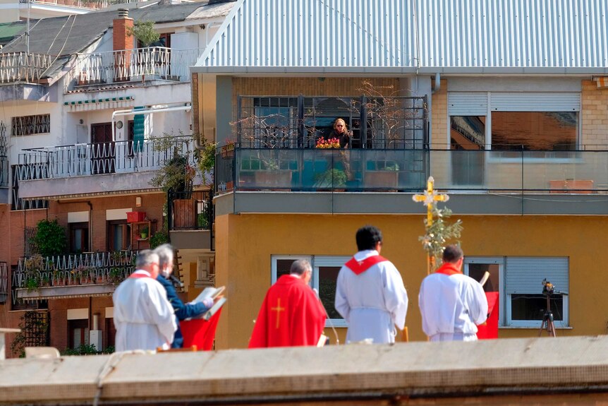 Priests say mass on a rooftop while a woman watches on from a balcony.