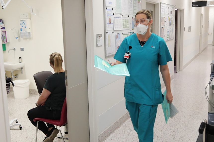 A female doctor in scrubs and a face mask walks down a hospital corrior