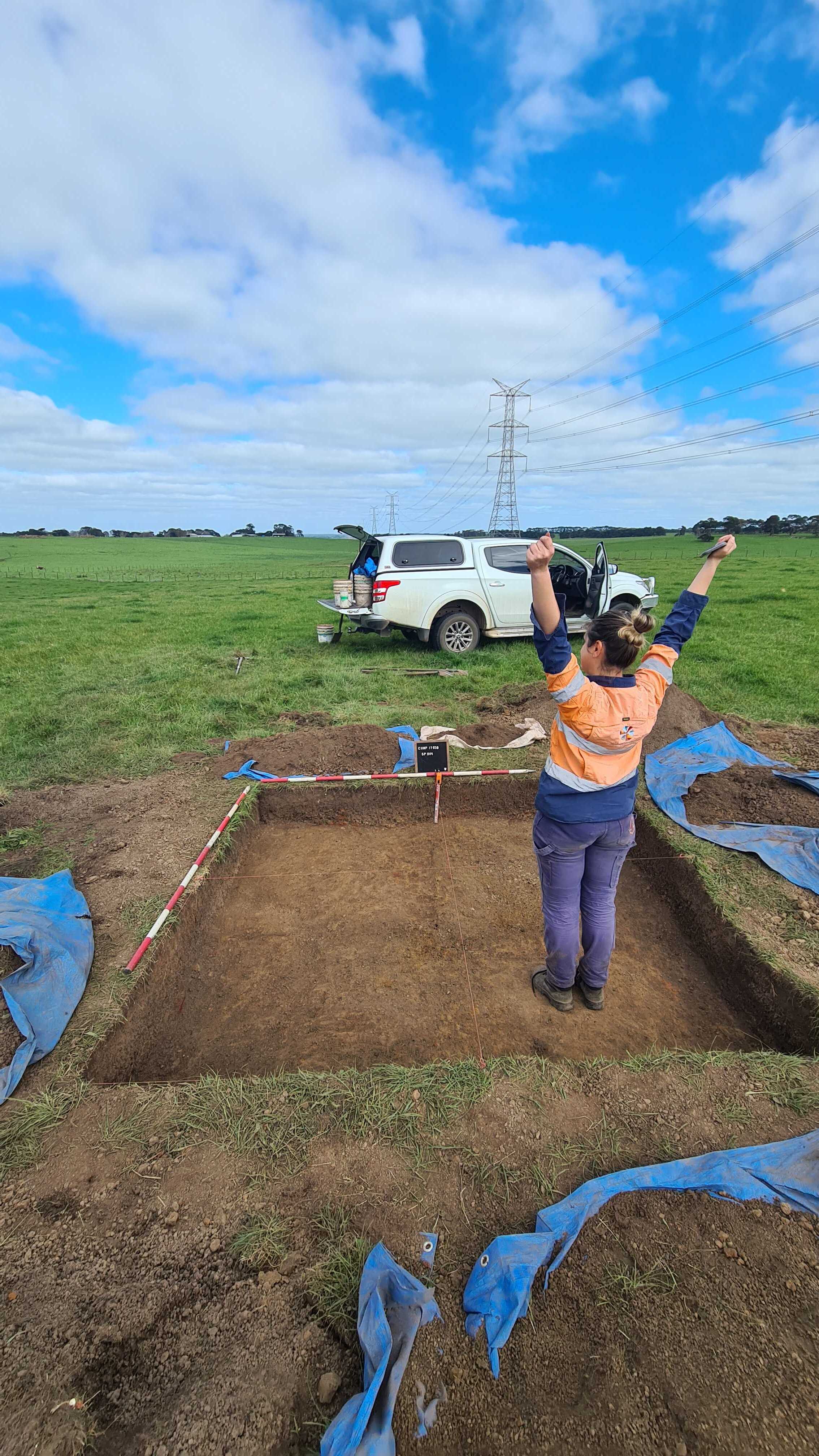 A woman in high vis stands in a dug out hole in a field with her arms up in celebration. 