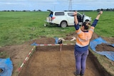 A woman in high vis stands in a dug out hole in a field with her arms up in celebration. 