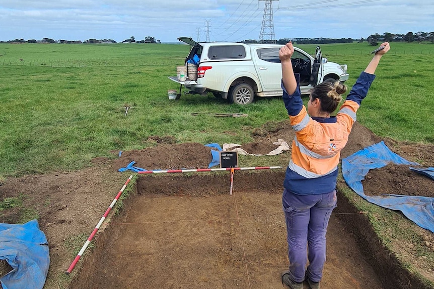 A woman in high vis stands in a dug out hole in a field with her arms up in celebration. 