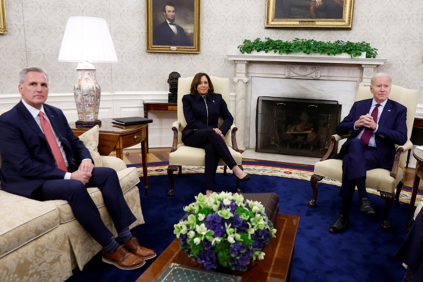 Two men and a woman sit in an ornate meeting room with paintings of former US presidents on the wall.