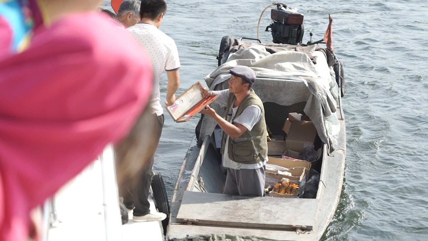 A man in a small boat shows off a souvenir to a larger boat full of tourists.