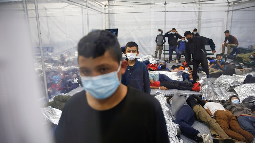 Children stand in masks next to others sleeping in a crowded space