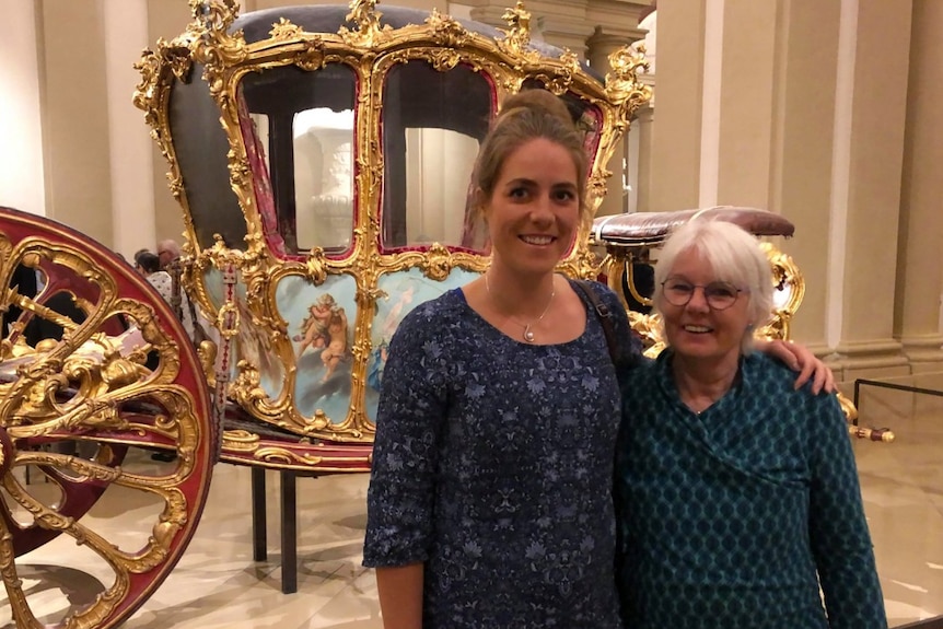 A young woman and an older woman pose in front of an opulent carriage.