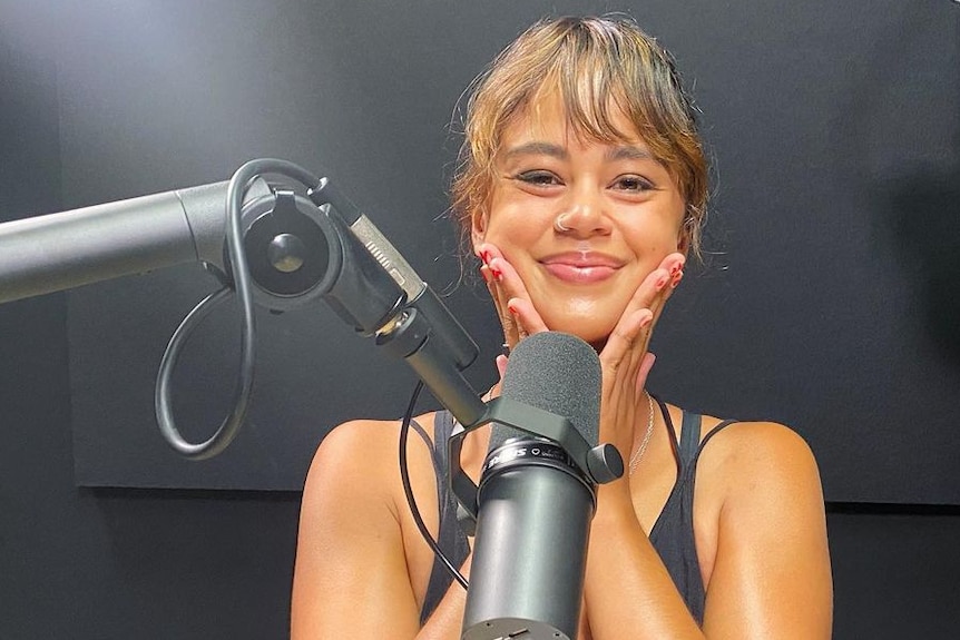 A brunette woman behind a podcast microphone smiling in a blackened studio 