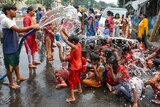 An adult hoses down a group of clothed children who splash the water as they sit on a street.