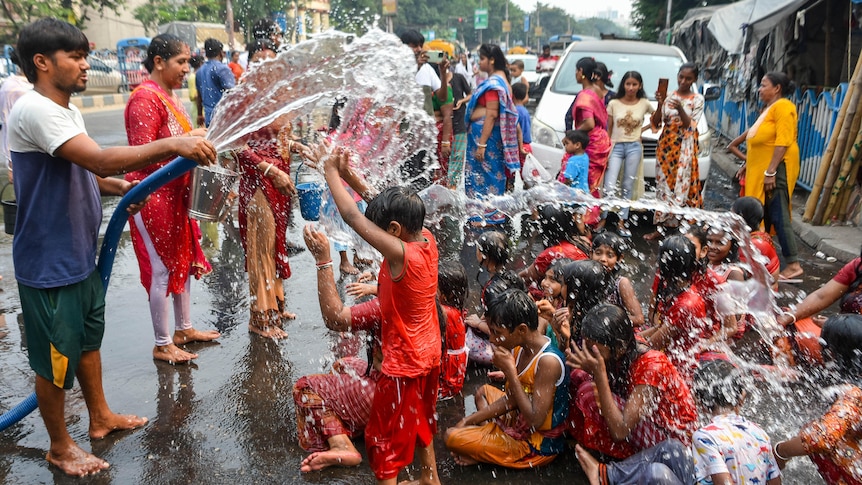 An adult hoses down a group of clothed children who splash the water as they sit on a street.