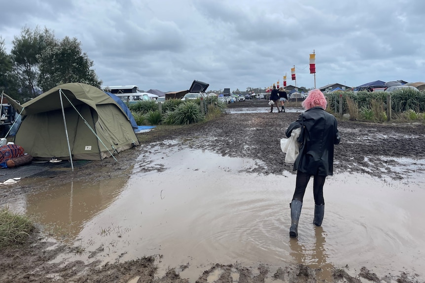 woman with pink hair wearing black clothes walks through puddles and mud, tent on the side