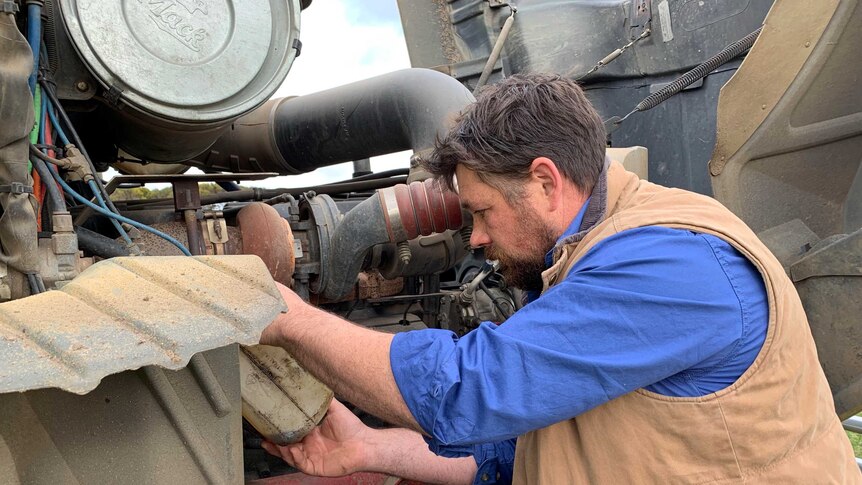 A man with brown hair and a scrubby beard working on a truck engine.