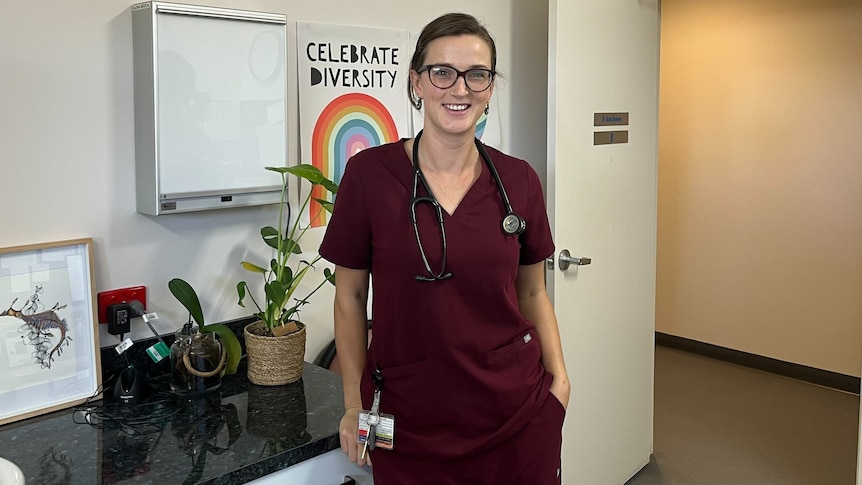 A woman wearing glasses and red scrubs stands against a counter with plants and artwork and a sign 'celebrate diversity'