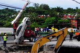 A crane lifts a car out of a large sinkhole, which opened on a highway. Rescue workers and other onlookers stand watching.