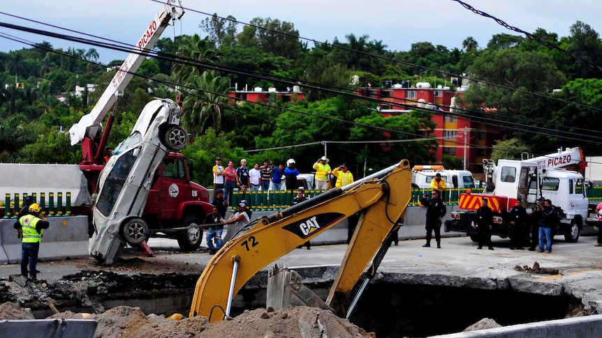 A crane lifts a car out of a large sinkhole, which opened on a highway. Rescue workers and other onlookers stand watching.