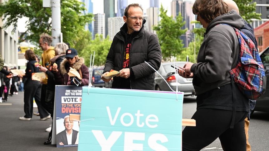Bandt, in a puffy jacket, stands on the street behind a "Vote Yes" sign, handing out flyers.