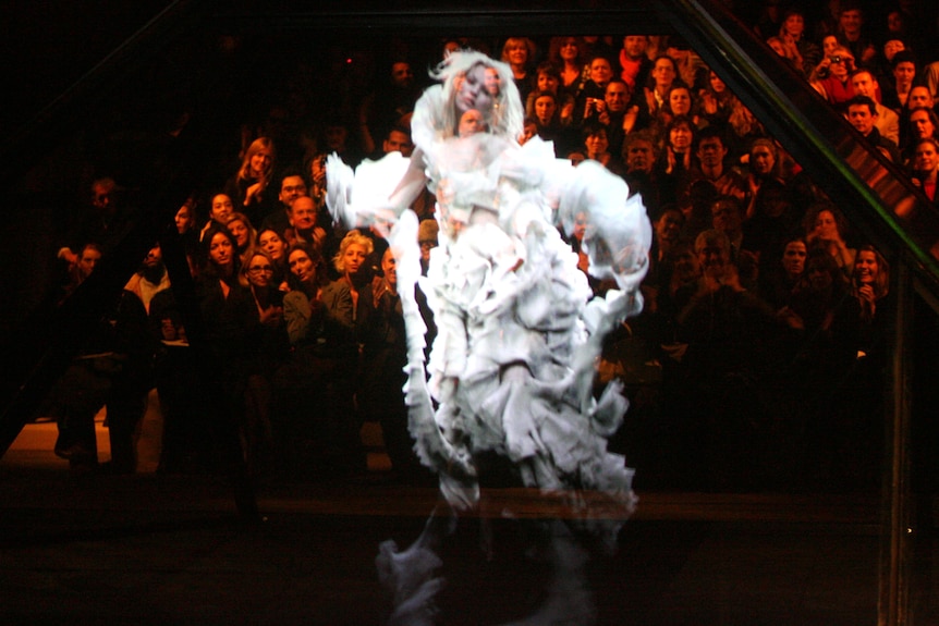 A hologram of model Kate Moss walks a catwalk in an extravagant white dress, with a crowd of people watching on