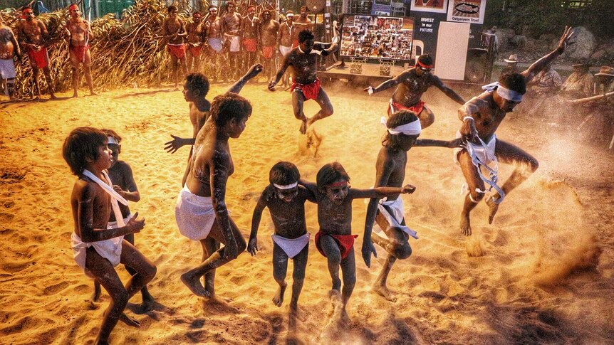 Indigenous adults and children kick up a cloud of sand as they perform a traditional dance.