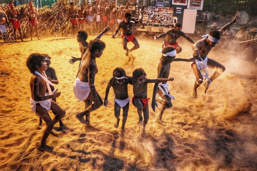 Indigenous adults and children kick up a cloud of sand as they perform a traditional dance.