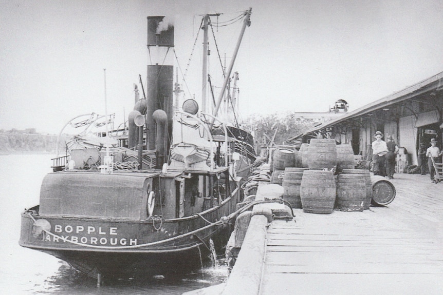 A black and white photo of a small ship moored at a wooden dock. 