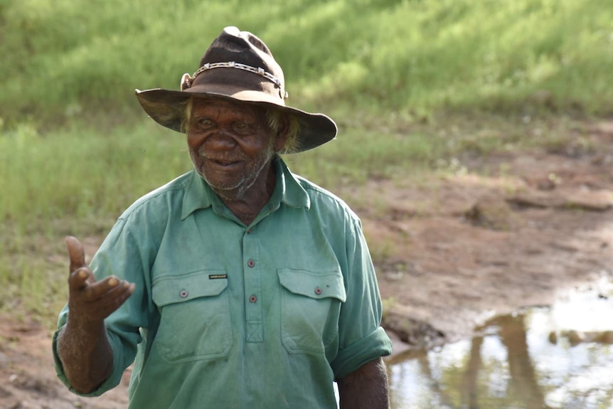 An Indigenous man in a hat stands in a remote rural setting.
