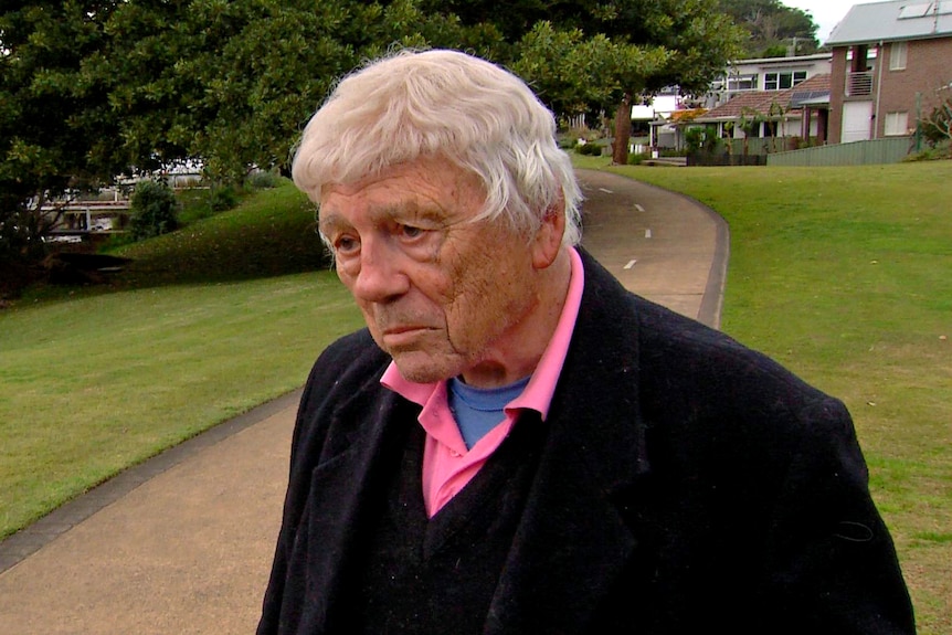 A man frowns standing on a concrete path through a grassed area. Some houses can be seen in the background.