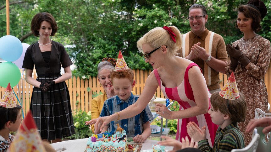 A child sits in front of a birthday cake outside surrounded by guests