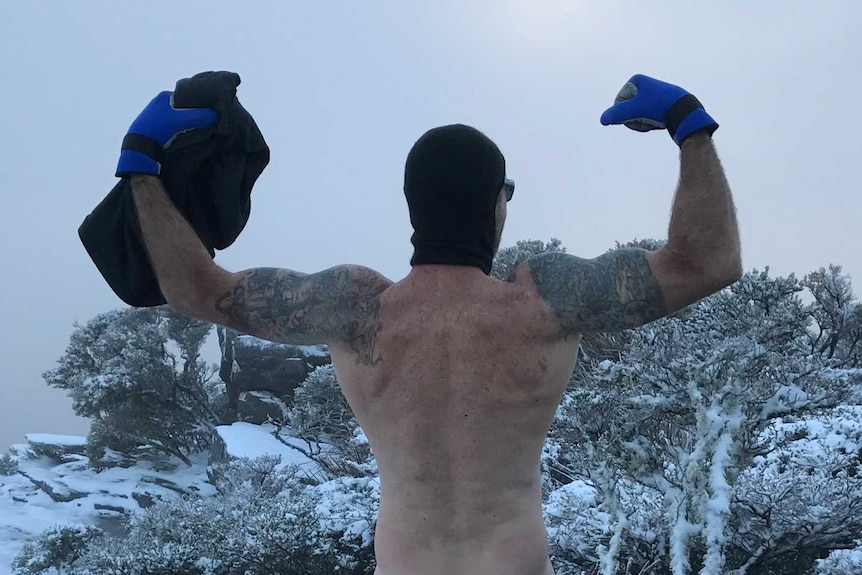Man stands naked on top of mountain surrounded by snow.