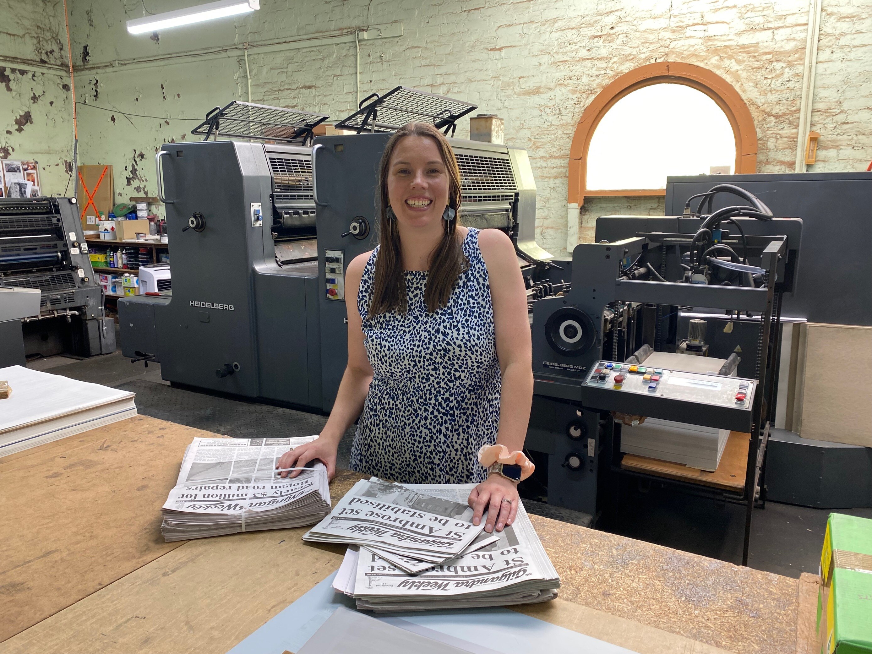 A woman stands in front of a newspaper printing press with her hands on two stacks of newspapers