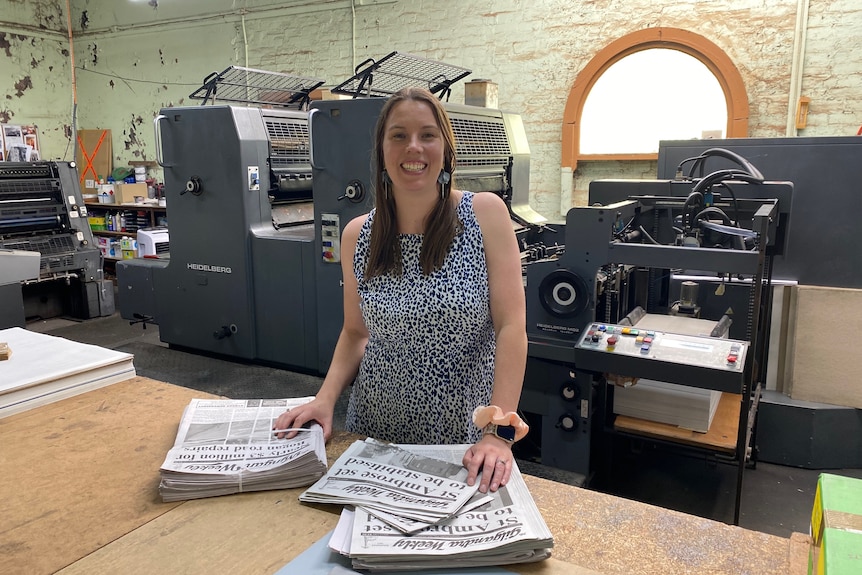 A woman stands in front of a newspaper printing press with her hands on two stacks of newspapers