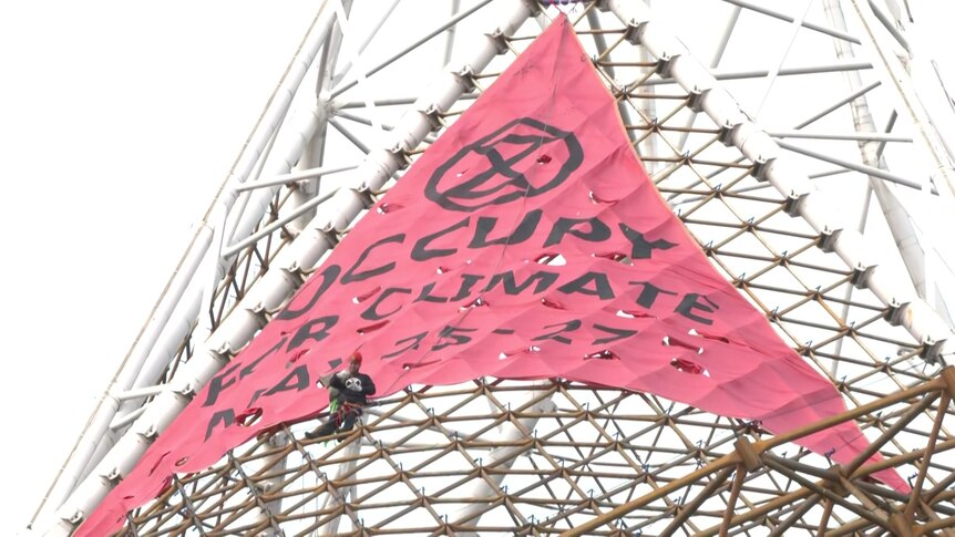 A man sits in front of a large pink banner that says 'occupy' on a spire