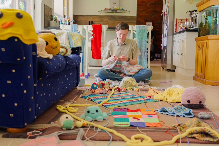 Jorden sits on his kitchen floor surrounded by 10s of completed crochet projects including animals. He crochets a blanket.