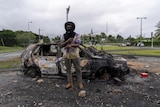 A man in a black hood stands in front of a burnt out car