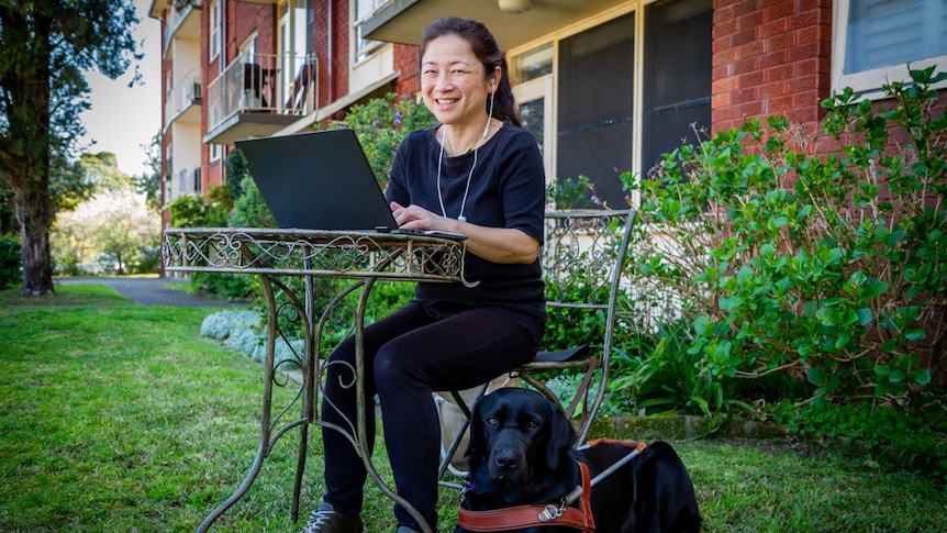 Lee Chong smiling while using a laptop on a small metal table outdoors, and a big black dog sits at her feet. 