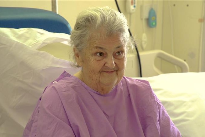 An elderly woman in a purple hospital gown sitting in a hospital bed