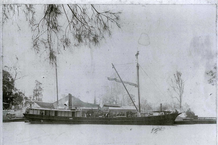 A black and white photo of a sailing ship in front of a timber mill on a river.