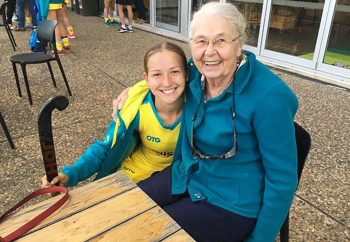 A young woman in an Australian hockey uniform kneeling next to her grandmother and holding a hockey stick.