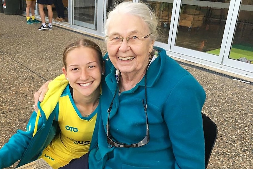A young woman in an Australian hockey uniform kneeling next to her grandmother and holding a hockey stick.