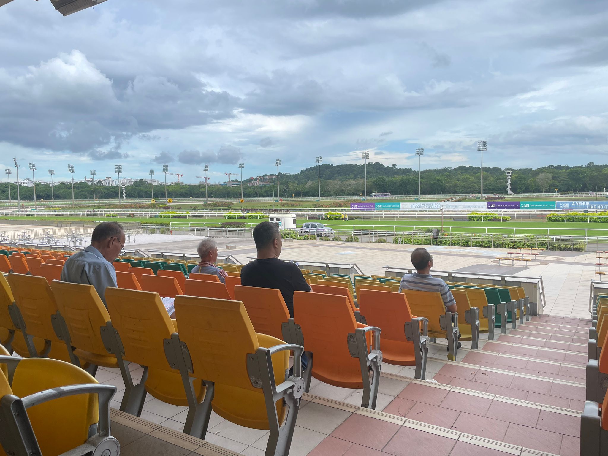 A few people sit on plastic seats in a large grandstand at a horse racing track.