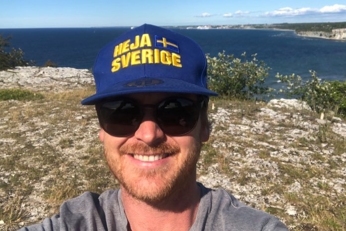A man taking a selfie at the edge of a cliff wearing a blue and yellow hat with the swedish flag.