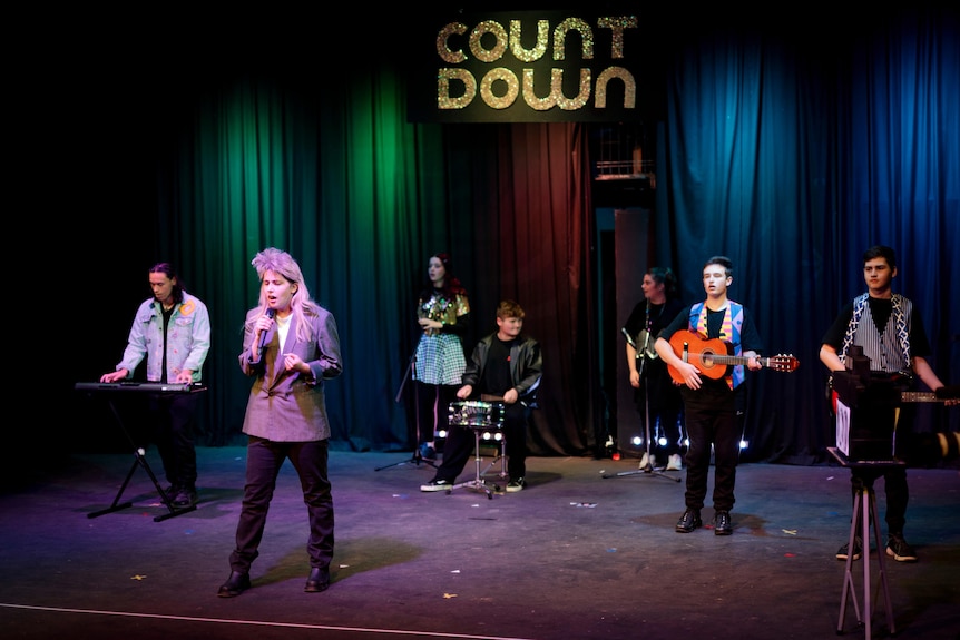 A teenage girl wearing a mullet wig sings on stage surrounded by a band. The sign Countdown hangs on the curtain