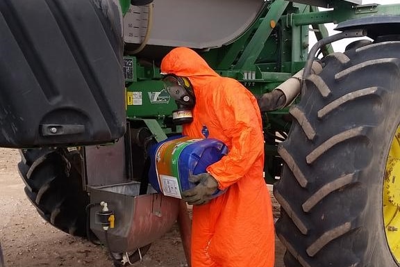 A man in a bright orange hazmat suit stand next to a piece of machinery unloading chemicals.