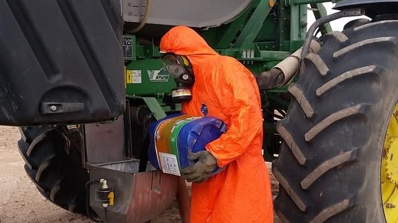 A man in a bright orange hazmat suit stand next to a piece of machinery unloading chemicals.