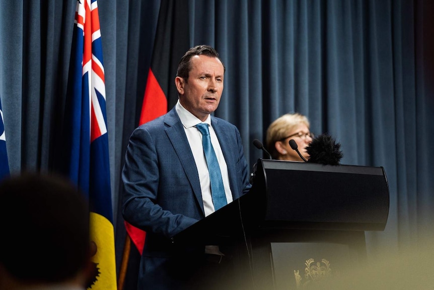 Mar McGowan wearing a blue suit, white shirt and light blue tie standing at a podium with microphones in front of flags.