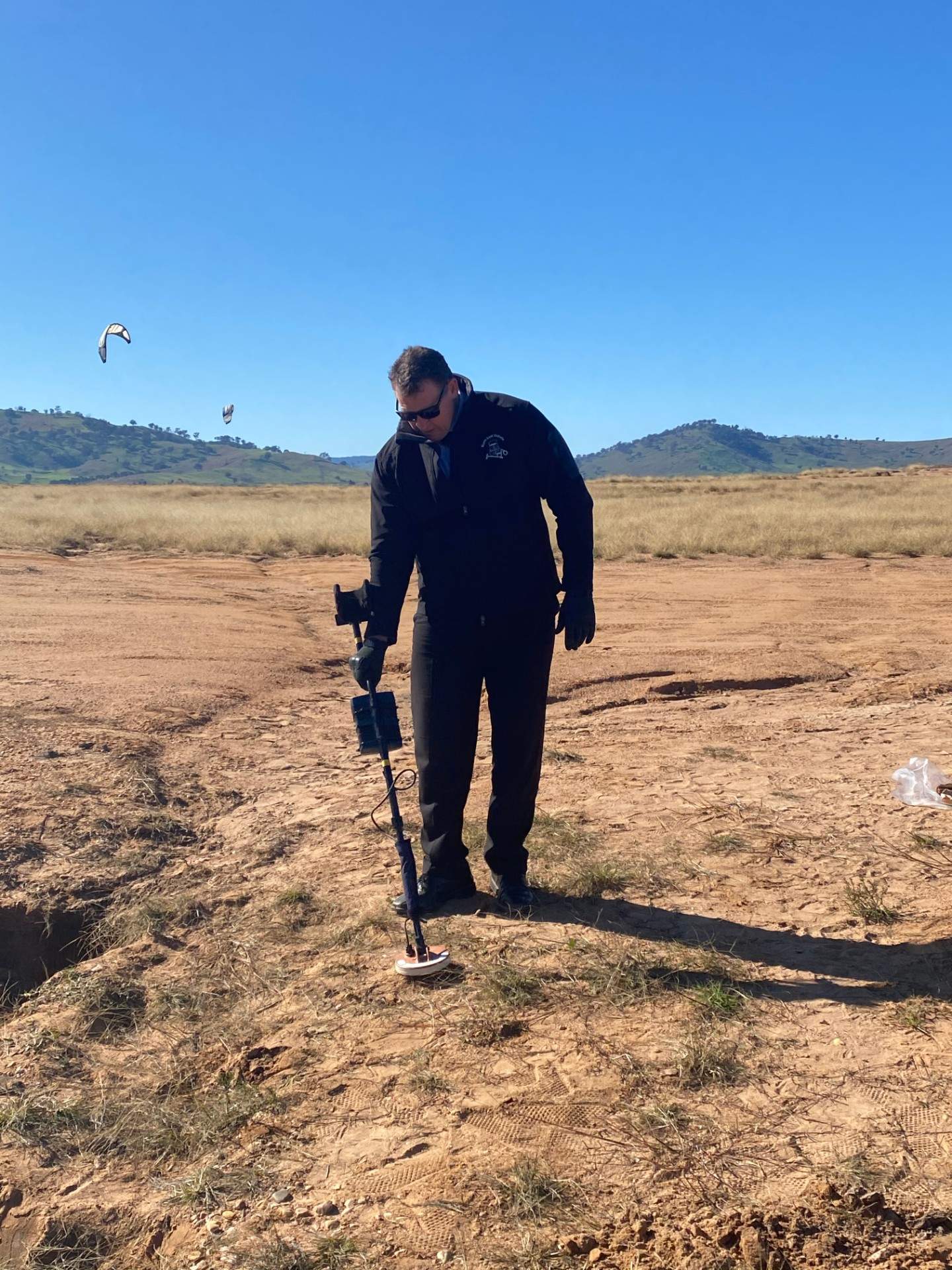 Man in dark clothing and sunglasses holds metal detector over desert like ground.