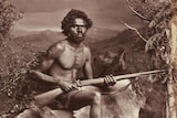 A sepia picture of an Indigenous man posing with a gun across his lap.
