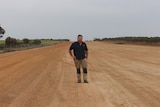 He stands on red dirt airstrip, with a hand on hip, navy shirt and cargo pants