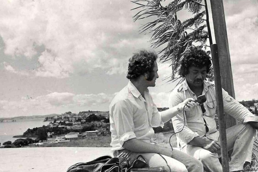 Black and white photo of a man holding a radio microphone interviewing a man sitting under a palm tree on a beach.