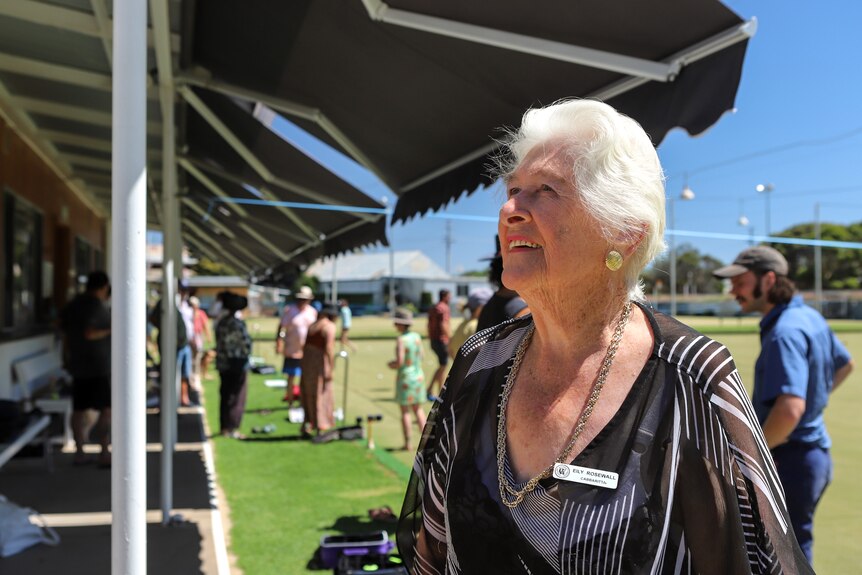 Woman with white hair wearing jewellery and blouse stands in front of lawn bowls field with people behind her