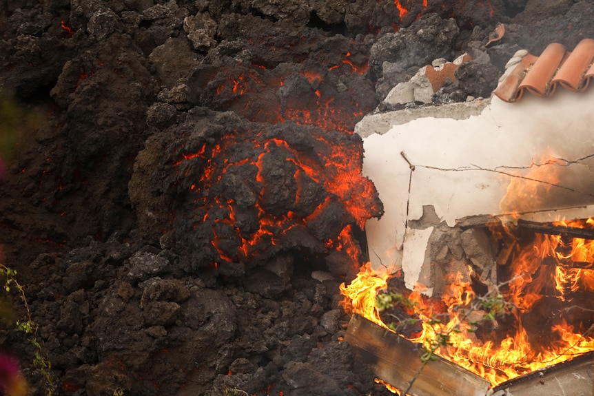A close-up of lava engulfing a wall. Just ahead of where the black magma touches it, the wall is on fire.