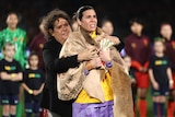 A woman soccer player is wrapped in a large cloak made of kangaroo skin by a famous Aboriginal sports icon