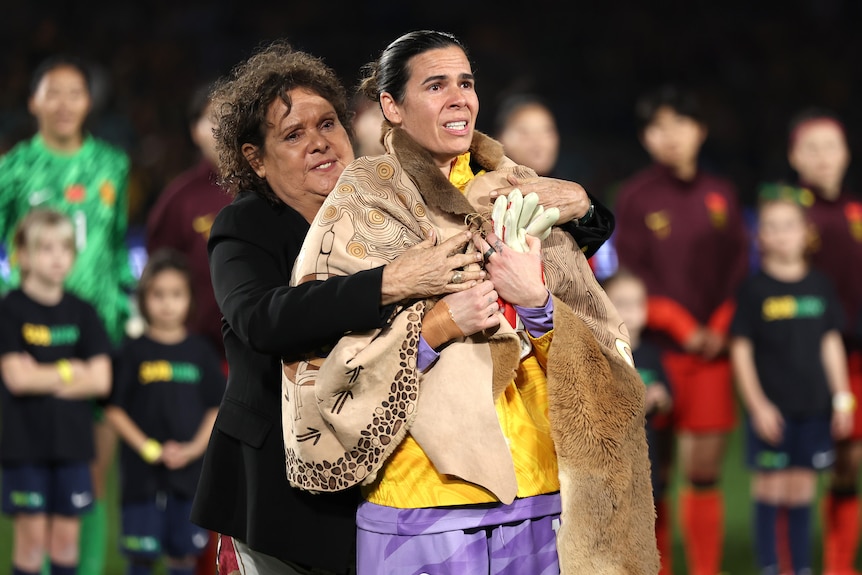 A woman soccer player is wrapped in a large cloak made of kangaroo skin by a famous Aboriginal sports icon